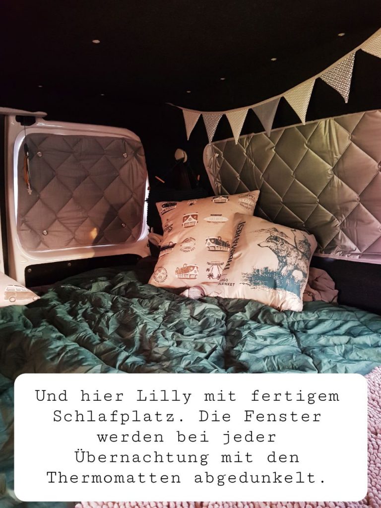 Blick auf die Schlafläche unseres Caddys. Ein Schlafsack auf einer Box, oben drauf ein paar Kissen fein arrangiert. Eine Wimpelkette hängt an der Kante zum Dach.